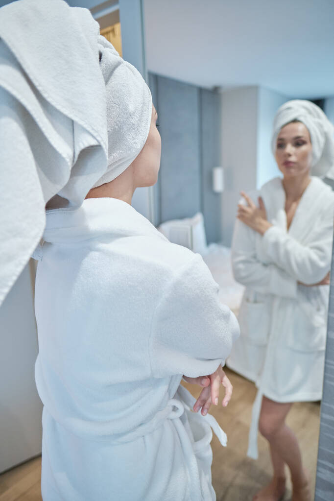 Είμαι όμορφη. Πορτρέτο της αυτοπεποίθηση νεαρή γυναίκα κοιτάζοντας τον καθρέφτη με ικανοποίηση, ενώ στέκεται στο μπουρνούζι στο σπίτι. Έννοια φροντίδας σώματος - Φωτογραφία, εικόνα