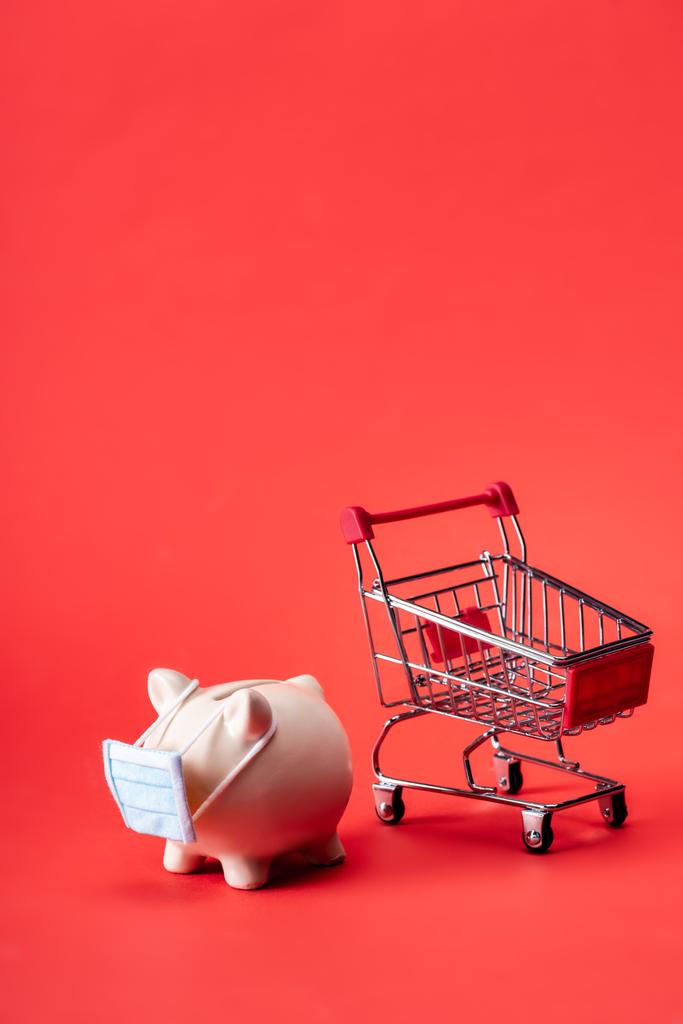 Sparschwein in kleiner medizinischer Maske neben Spielzeug-Einkaufswagen auf Rot  - Foto, Bild