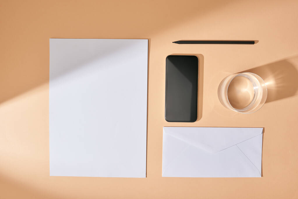 ベージュの背景に紙、スマートフォン、鉛筆、水と封筒のシートでフラットレイ - 写真・画像