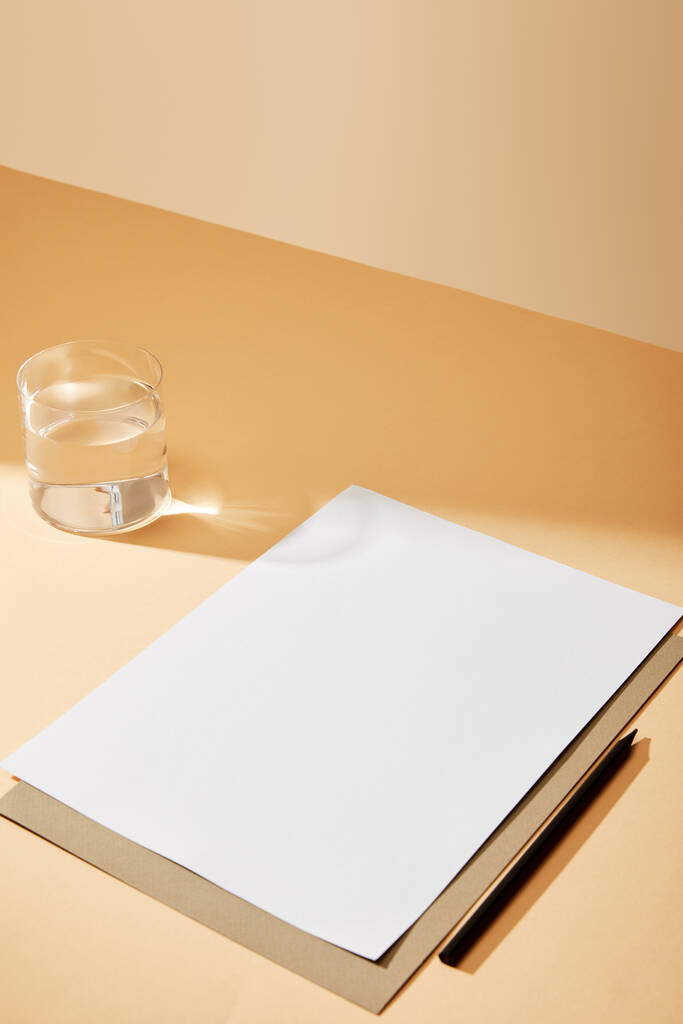 φύλλο χαρτιού και μολυβιού κοντά σε γυαλί νερού σε μπεζ επιφάνεια - Φωτογραφία, εικόνα