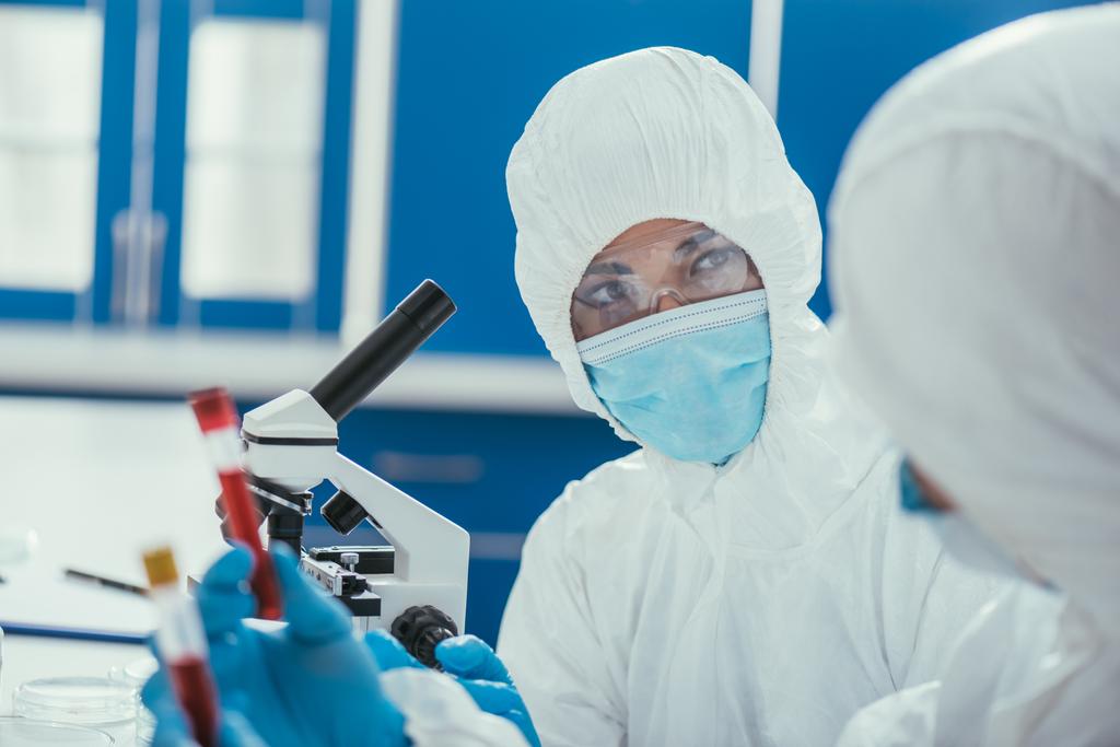 biokémikus vegyvédelmi ruhában nézi kollégáját, aki vérmintákkal ellátott kémcsöveket tart - Fotó, kép