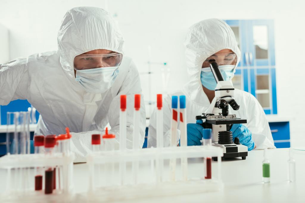biochemik pracující s mikroskopem v blízkosti kolegy při pohledu na zkumavky v laboratoři - Fotografie, Obrázek