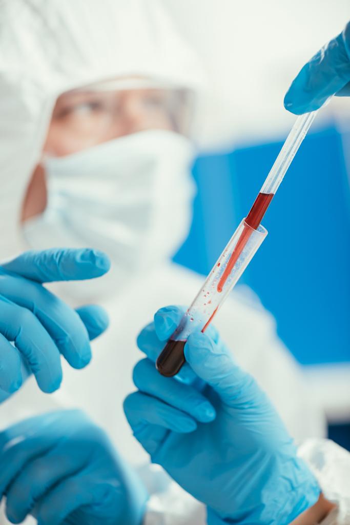 vista recortada del bioquímico sosteniendo la pipeta y el tubo de ensayo con muestra de sangre cerca de su colega
 - Foto, Imagen