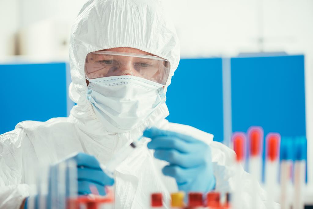 Tehlikeli madde giysisi içindeki biyokimyacı laboratuarda test tüplerinin yanında şırınga tutuyor. - Fotoğraf, Görsel
