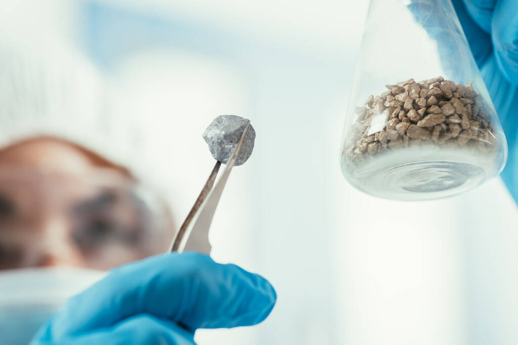 selective focus of biochemist holding small stone with tweezer and flask with gravel - Photo, Image