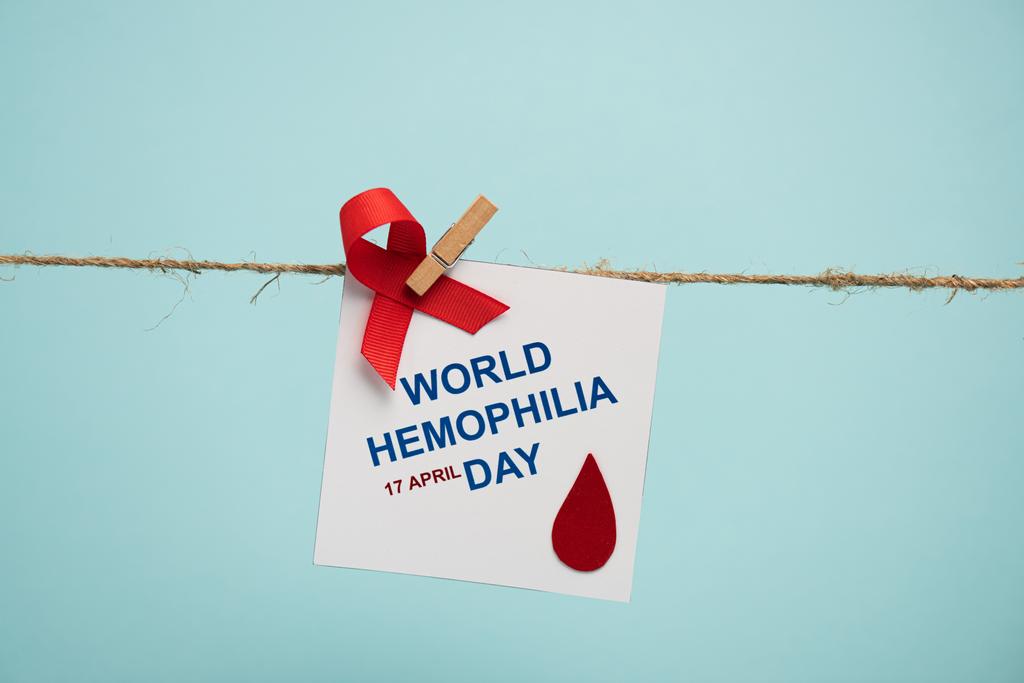 Karte mit Schriftzug zum Welttag der Hämophilie und rotem Band auf Seil mit Nadel isoliert auf blau  - Foto, Bild
