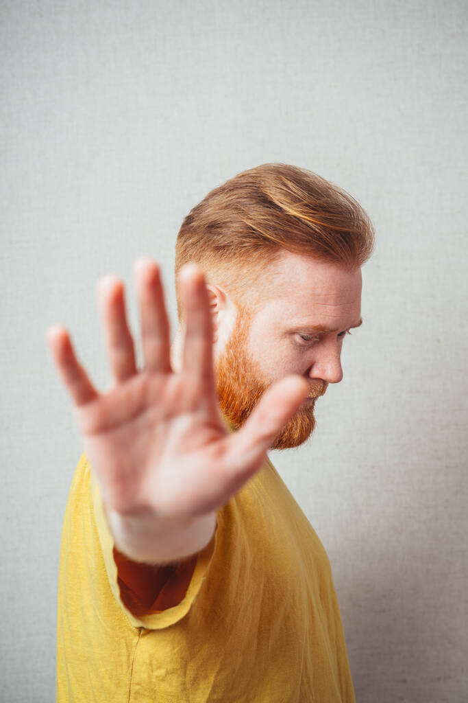 Sarı tişörtlü, sakallı, gri arka planlı bir adam öfkeden kuduruyor ve durduğunu gösteriyor. - Fotoğraf, Görsel