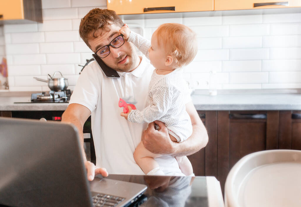 Όμορφος νεαρός που εργάζεται στο σπίτι με ένα φορητό υπολογιστή με ένα μωρό στα χέρια του. Μείνε σπίτι. Κεντρικό γραφείο με παιδιά.  - Φωτογραφία, εικόνα