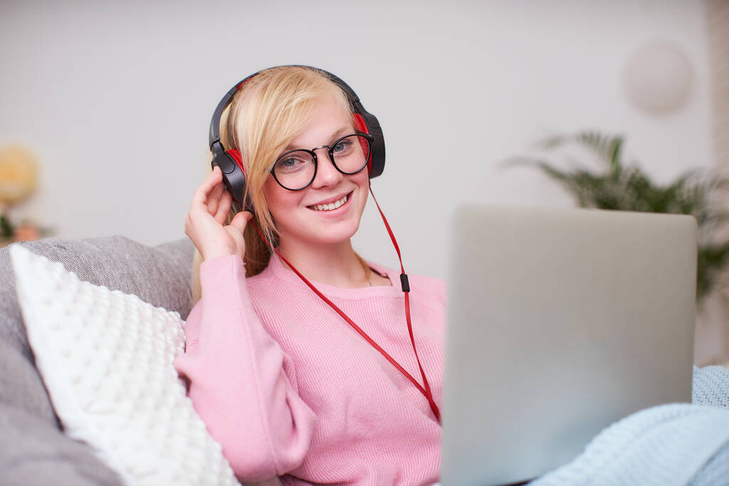 女の子の学生は、インターネットを介して友達のヘッドフォンと通信して幸せ。家庭教育仕事勉強新しい知識。幸せなティーンはコンピュータと椅子に座っています。女性はノートパソコンを見て笑顔で. - 写真・画像