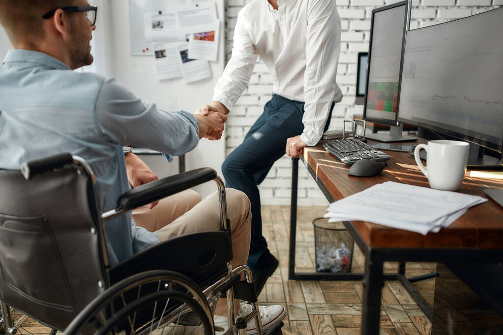 Tanıştığıma memnun oldum. Yaratıcı ofiste birlikte çalışırken tekerlekli sandalyede iş arkadaşına el sıkışan genç bir ofis çalışanı. Engelli konsepti - Fotoğraf, Görsel