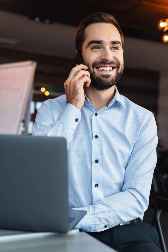 Portrét pohledného mladého podnikatele v bílé košili mluvící na mobilu a pomocí notebooku při práci v kanceláři - Fotografie, Obrázek