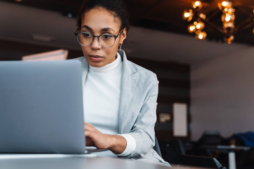 Beeld van jonge brunette Afrikaans Amerikaanse zakenvrouw zitten aan tafel en werken op laptop in kantoor kamer - Foto, afbeelding