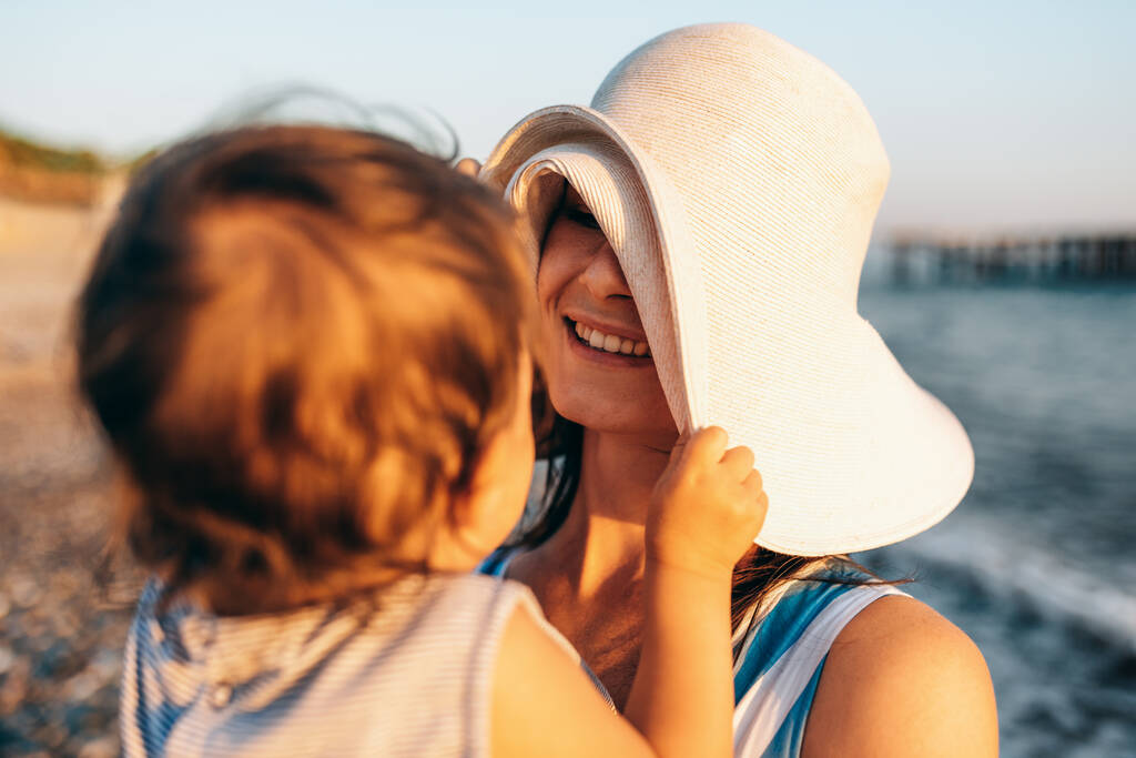 Mutlu gülümseyen genç anne ve küçük kızının portresi deniz batımında eğleniyor. Güzel bir dişi ve bebek gün batımında okyanus sahilinde birlikte oynuyorlar. Annelik aşkı bakımı. - Fotoğraf, Görsel
