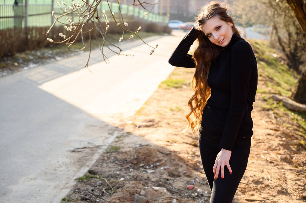 Портрет красивой молодой девушки с длинными волосами с улыбкой на фоне дорожных деревьев в парке. Фото в солнечную погоду на открытом воздухе весной
. - Фото, изображение