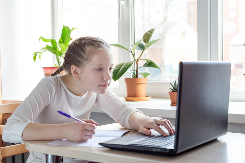 Koulutyttö katsoo kannettavan tietokoneen näyttöä. Sosiaalinen etäisyys, eristäytyminen. Kotikoulu, verkko-opetus, kotiopetus, karanteenikäsittely - Kuva - Valokuva, kuva