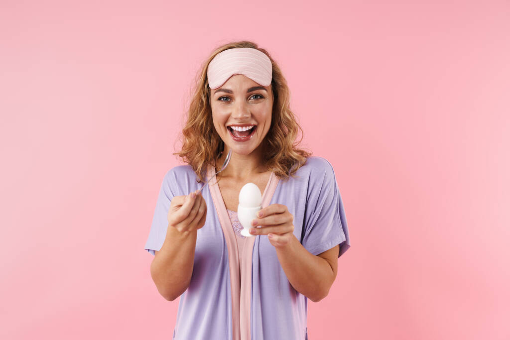 Imagem da jovem mulher excitada em máscara de sono sorrindo enquanto come ovo isolado sobre fundo rosa
 - Foto, Imagem
