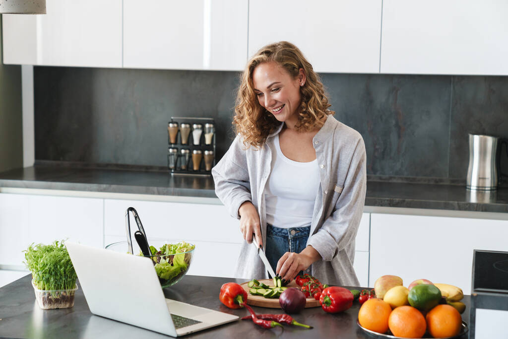 Szczęśliwa młoda kobieta robi sałatkę w kuchni, sieka warzywa, patrzy na laptopa - Zdjęcie, obraz