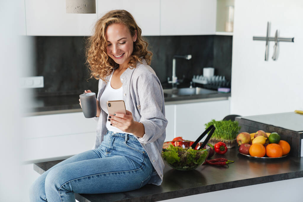 Glimlachende jonge vrouw in casual kleding met een kopje koffie terwijl ze op een keukentafel zit en mobiele telefoon gebruikt - Foto, afbeelding