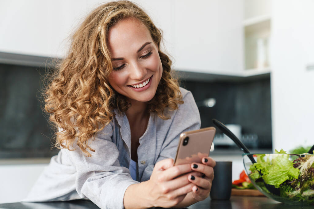 Mujer joven sonriente usando ropa casual usando teléfono móvil mientras se apoya en una mesa de cocina
 - Foto, imagen