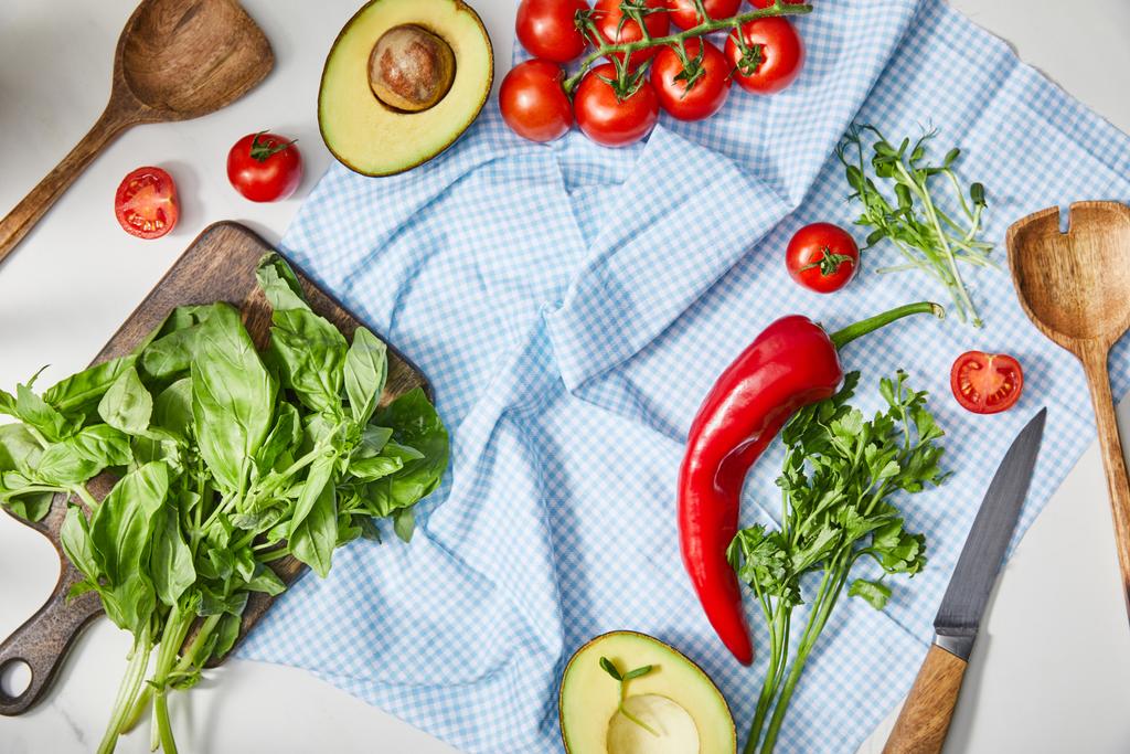 Vista superior de verduras, vegetación, mitades de aguacate, cuchillo y espátulas en tela a cuadros cerca de albahaca en tabla de cortar en blanco
  - Foto, imagen