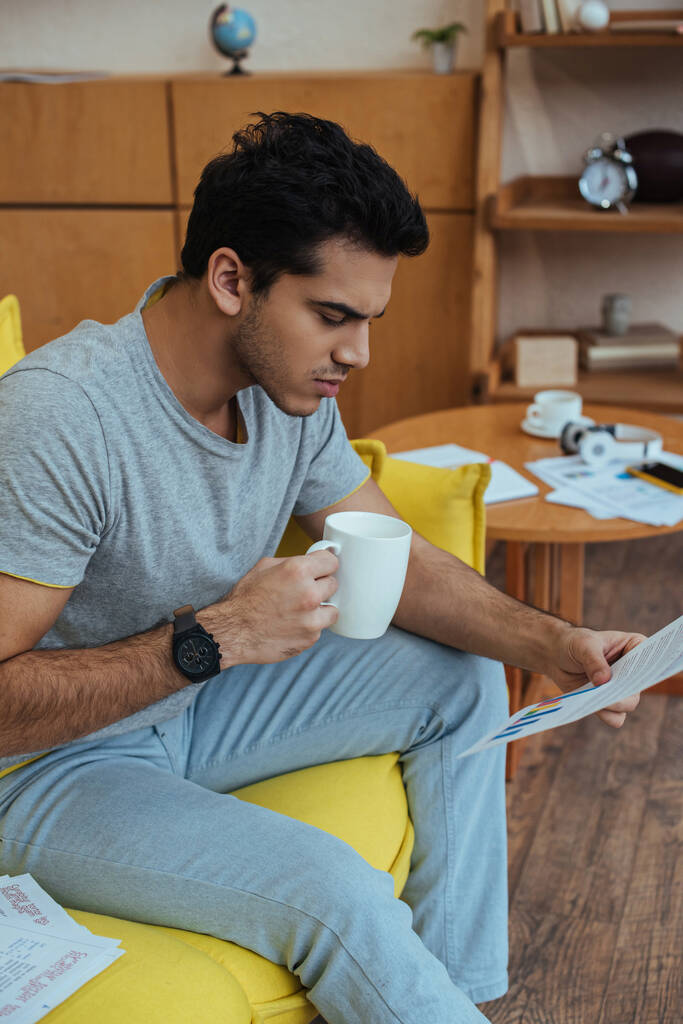 Freelancer con taza de té mirando el papel y sentado en el sofá en la sala de estar
 - Foto, imagen