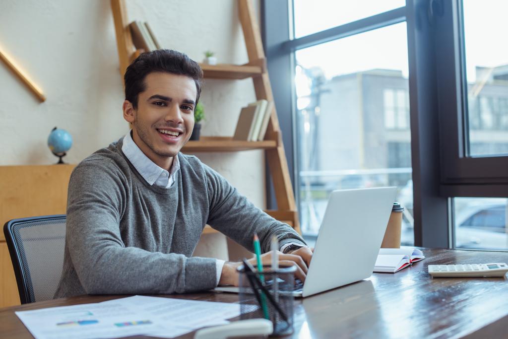 Селективный фокус бизнесмена, работающего с ноутбуком, улыбающегося и смотрящего на камеру за столом в офисе
 - Фото, изображение