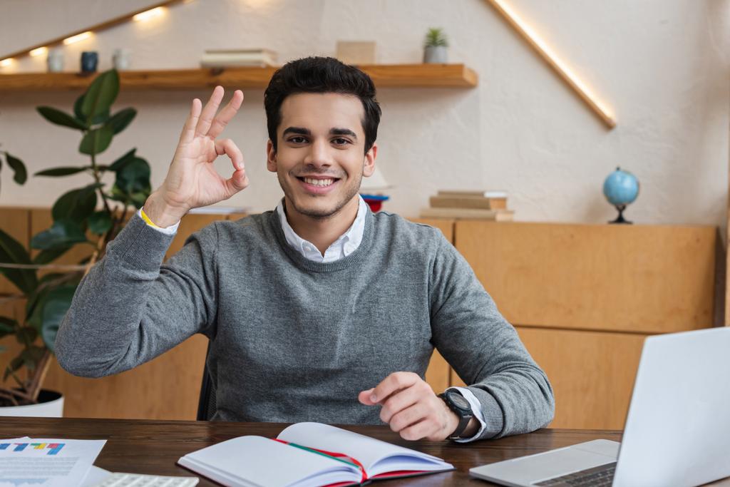Businessman looking at camera, smiling and showing okay sign at table in office - Photo, Image
