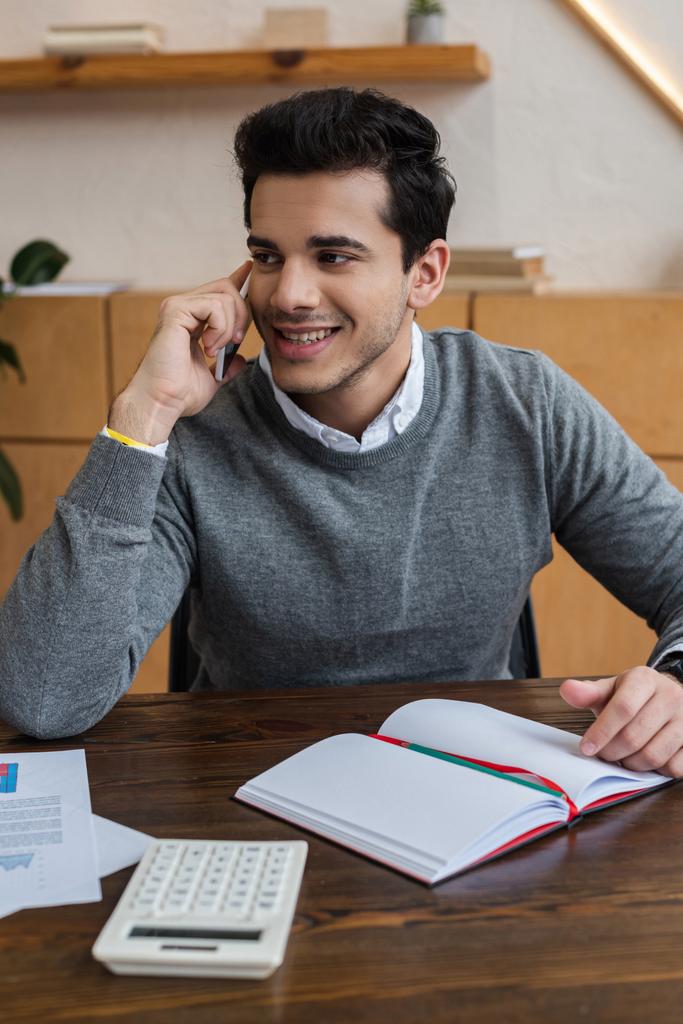 Podnikatel se usmívá, dívá se jinam a mluví na smartphone u stolu v kanceláři - Fotografie, Obrázek