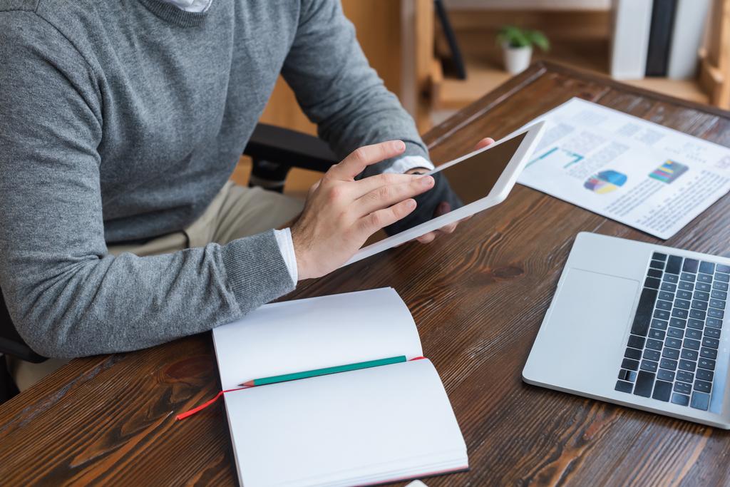 Ausgeschnittene Ansicht eines Geschäftsmannes, der mit digitalem Tablet in der Nähe von Notebook und Laptop am Tisch im Büro arbeitet - Foto, Bild