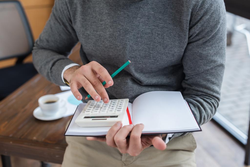 Vista recortada de hombre de negocios con lápiz y cuaderno usando calculadora en la oficina
 - Foto, Imagen