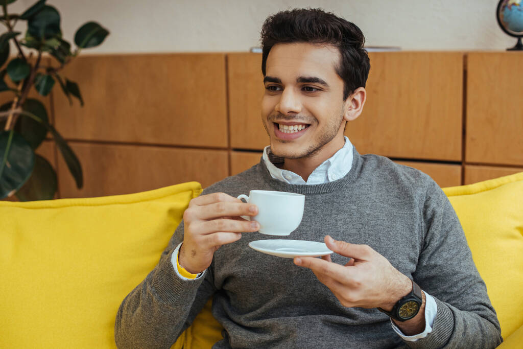Muž s úsměvem a podšálkem a šálkem kávy na pohovce - Fotografie, Obrázek
