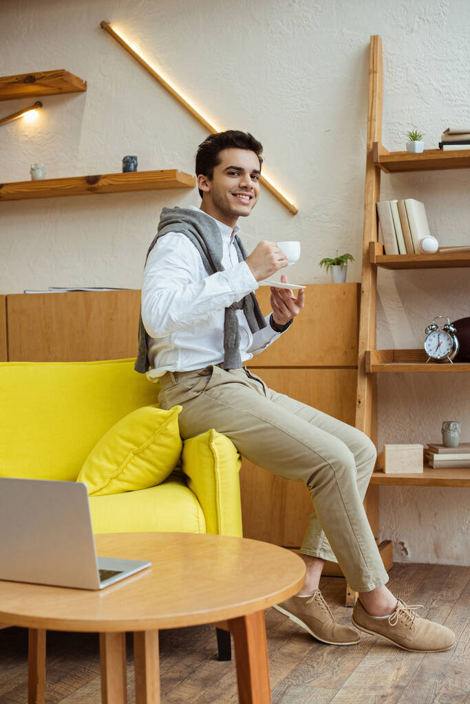Hombre de negocios con taza de café sonriendo y mirando a la cámara en el sofá cerca de la mesa de café con el ordenador portátil en la oficina
 - Foto, Imagen