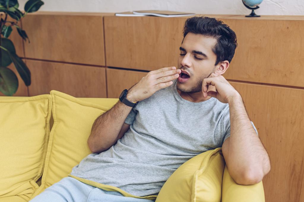 Человек зевает и покрывает рот на диване в гостиной
 - Фото, изображение
