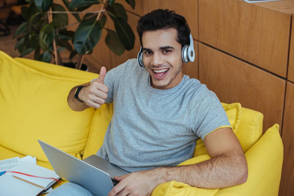 Vista de alto ángulo del freelancer en auriculares con portátil sonriendo, mirando a la cámara y mostrando como signo en el sofá
 - Foto, Imagen