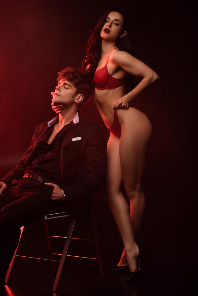 passionate girlfriend in red lingerie hugging sexy boyfriend in suit on black with red light - Photo, Image
