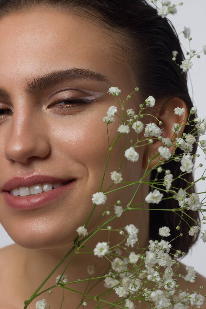 Close up portrait of a face with beautiful skin and nude makeup, eyeliner and long eyelashes, puffy natural lips and white flowers. Fashion photography. Cosmetology and spa. Freshness of spring - Photo, Image