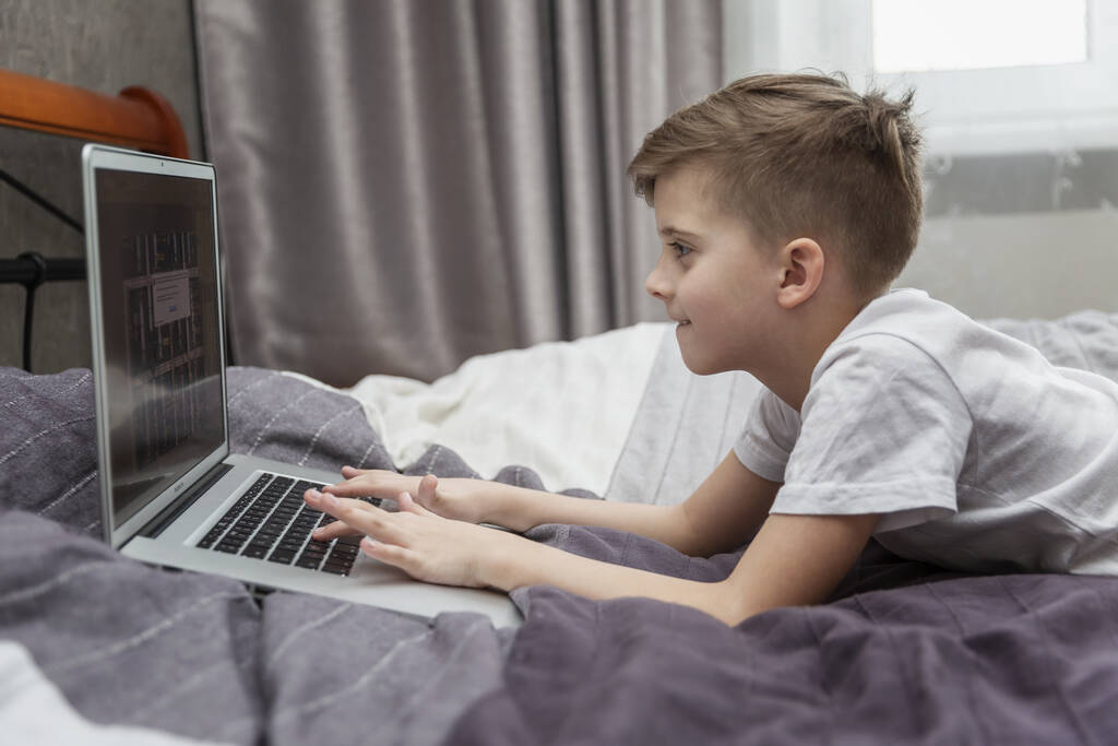 Un chico frente a un portátil en casa en la cama. Enseñanza a distancia. Cuarentena en el contexto de una pandemia de coronavirus
. - Foto, imagen