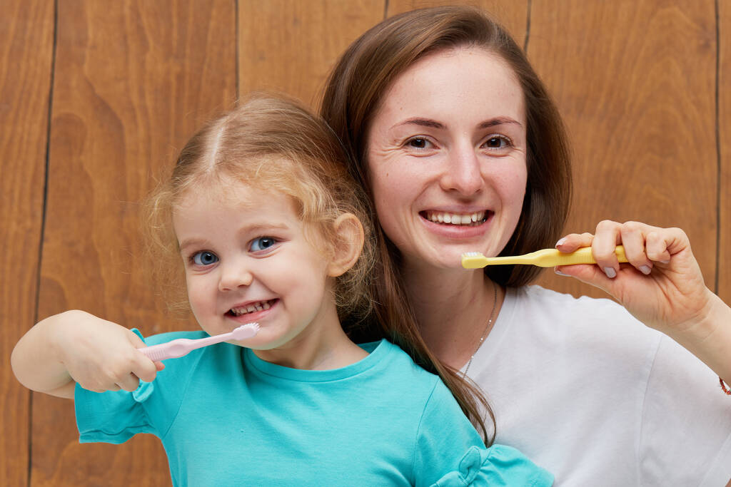 Higiene oral. Niña con madre cepillándose los dientes con un cepillo de dientes y mirando a la cámara sobre fondo de madera
 - Foto, Imagen