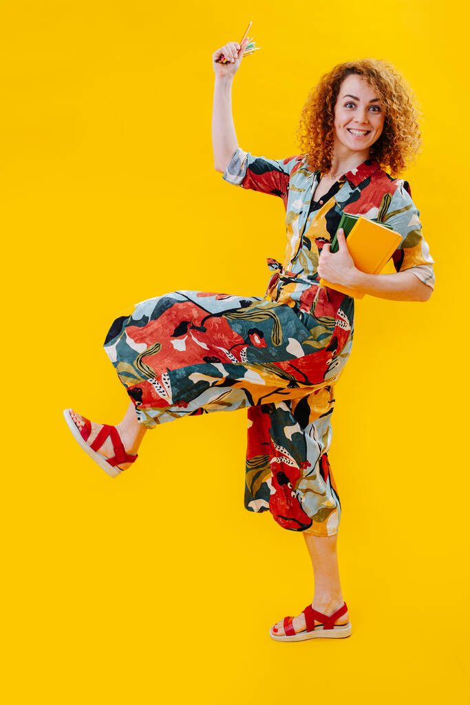 menina feliz andar levantando uma perna alta, em macacão colorido, segura um bloco de notas e lápis em suas mãos, ela tem cabelo encaracolado vermelho, imagem em um fundo amarelo
 - Foto, Imagem