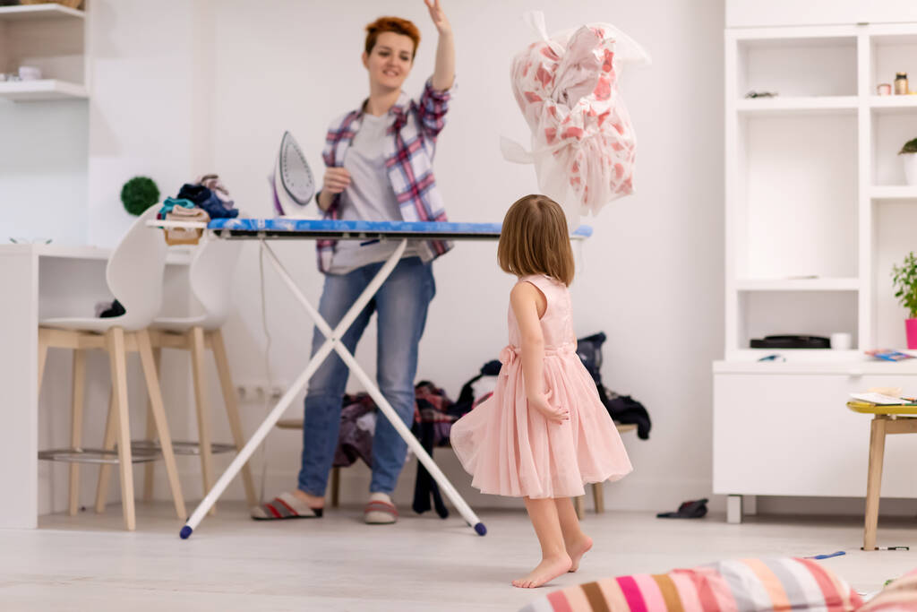 glückliche Familie hat Spaß zusammen zu Hause niedliche kleine Tochter in einem rosa Kleid spielen und tanzen, während junge rothaarige Mutter bügelt Kleidung hinter ihr - Foto, Bild