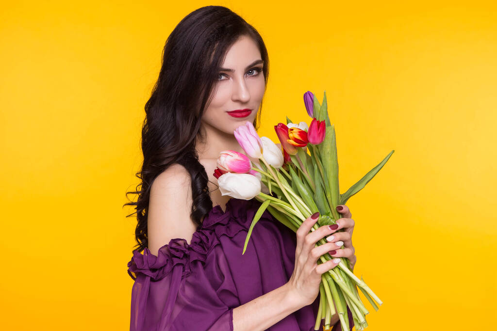 Красивая брюнетка с букетом тюльпанов в руках на жёлтом фоне. Женщина одета в фиолетовое, тонкое платье. 8 марта, день матри
. - Фото, изображение