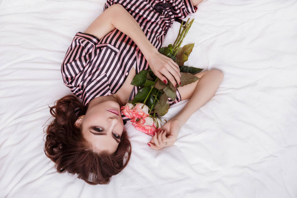 Mooi sexy, sexy meisje in gestreepte satijnen pyjama ligt op een wit bed met rozen. Een cadeau voor 8 maart, voor moederdag. - Foto, afbeelding