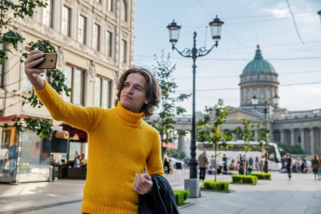 Οι όμορφοι άνδρες κάνει selfie, Κοιτάξτε από έξω, αυτός ντυμένος με ένα κίτρινο πουλόβερ, ένα μαύρο αδιάβροχο ή σακάκι είναι τα χέρια του, Bolshaya Konyushennaya δρόμο και Kazan καθεδρικό ναό στο παρασκήνιο, ηλιόλουστη μέρα - Φωτογραφία, εικόνα