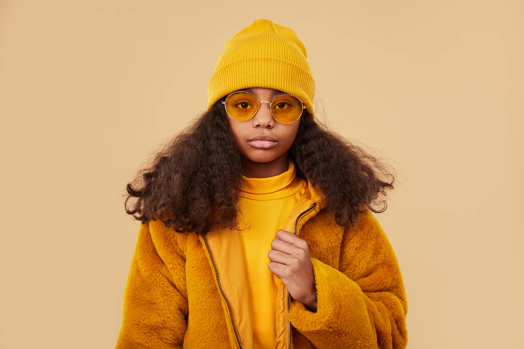 Moderna chica adolescente afroamericana confiada en gafas de moda con abrigo amarillo y suéter con sombrero de punto mirando a la cámara en fondo beige
 - Foto, Imagen