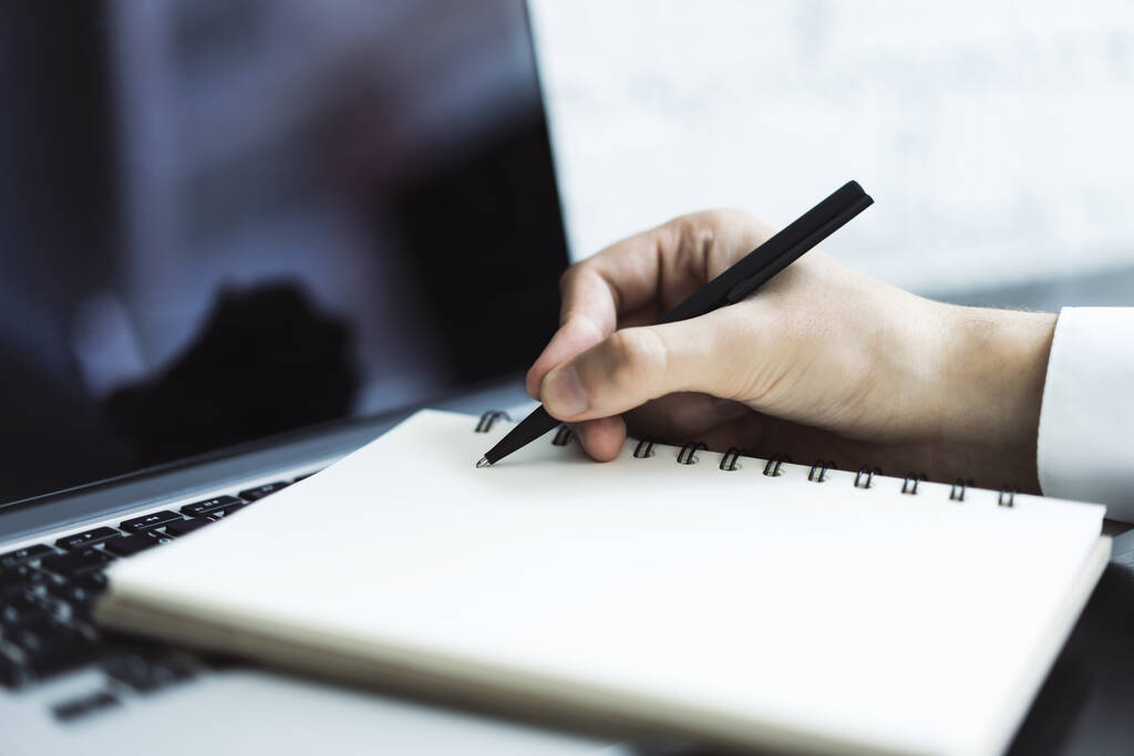 Man schrijft met een pen in notitieboekje op laptop toetsenbord in een zonnig kantoor-, bedrijfs- en onderwijsconcept. Sluiten. - Foto, afbeelding