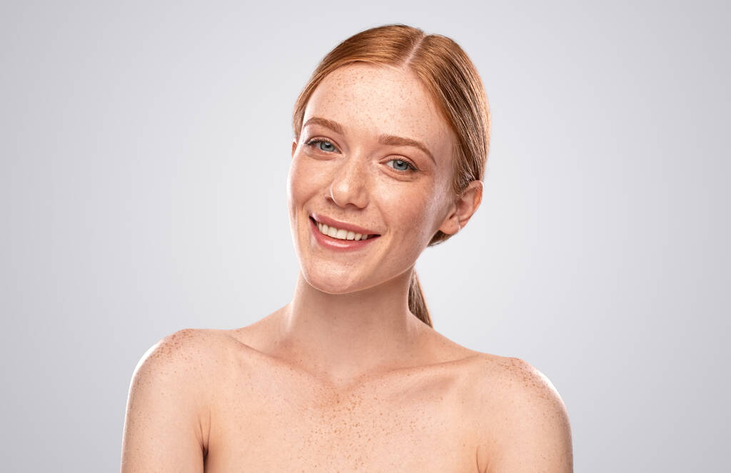 Atractiva joven hembra con el pelo rojo y la piel pecosa mirando a la cámara y sonriendo mientras representa la industria de la belleza contra el fondo gris
 - Foto, imagen