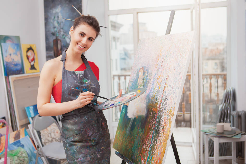 Aantrekkelijke vrolijke jonge vrouw die geniet van het schilderen in de kunststudio. Vrolijke vrouwelijke kunstenaar tekening in workshop - Foto, afbeelding