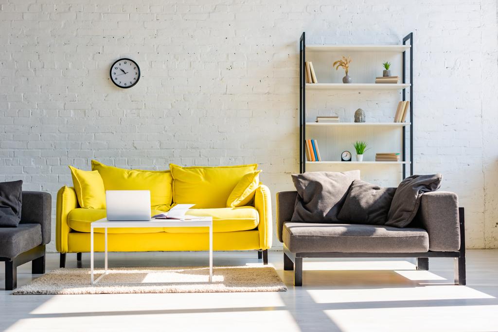 Wohnzimmer mit gelbem Sofa, grauen Sesseln, Regal, Uhr und Laptop im Sonnenlicht - Foto, Bild