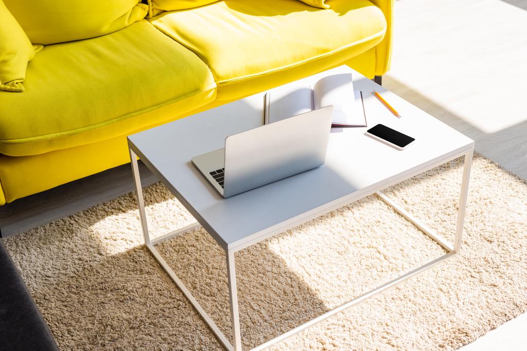 гостиная с желтым диваном и столом с ноутбуком, смартфоном и блокнотом в солнечном свете
 - Фото, изображение