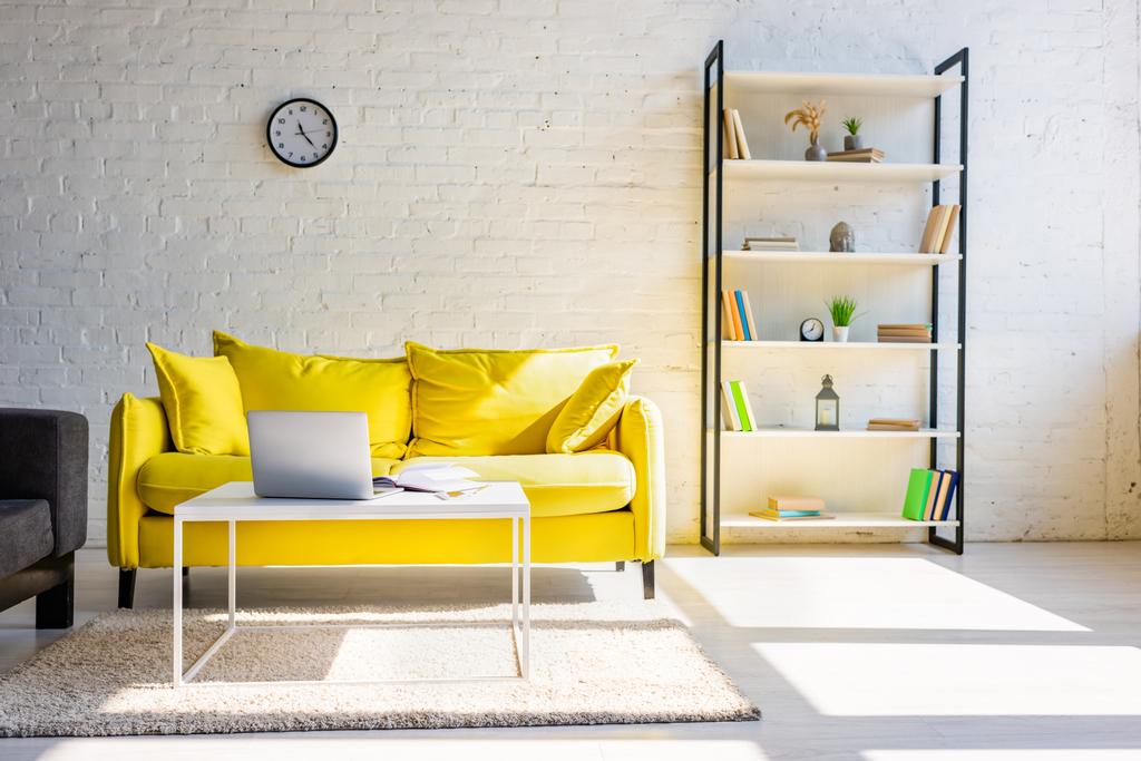 nappali sárga kanapéval, polc és asztal laptoppal napfényben - Fotó, kép